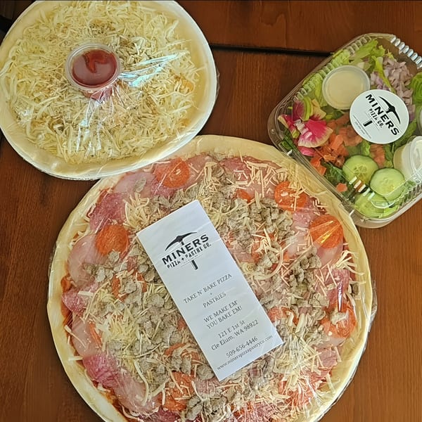 Two Miners Pizza Co. take-and-bake pizzas and a side salad with dressing on a wooden table.