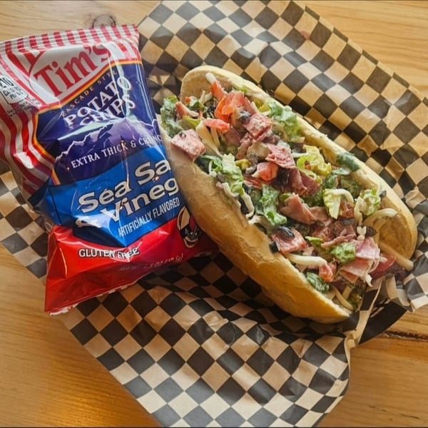 A loaded sub sandwich with lettuce, meat, and olives, next to a bag of Tim's Sea Salt & Vinegar chips.